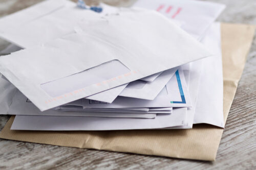A pile of various white envelopes, including some with windows, lies scattered on a wooden surface. One brown envelope is partially visible underneath the pile—perhaps containing news from the U.S. consulate about a visa denial.