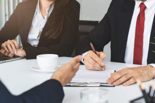 Three people in business attire sit at a table; one is signing a document, possibly related to citizenship test consequences, while another holds a clipboard. A white coffee cup sits on the table. Only their hands and torsos are visible.